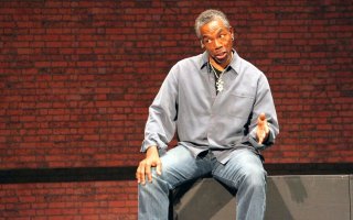 A seated man in jeans and a gray shirt gestures while speaking, his head tilted to one side.