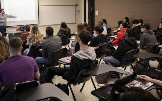 Man in gray sweater points to a projection as he speaks to a classroom full of students.