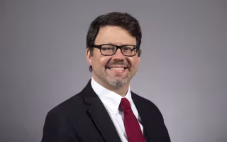 Portrait of a smiling man with short brown hair and glasses, wearing a suit and tie against a gray backdrop.
