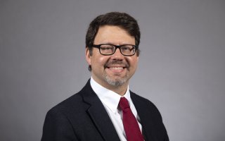 Portrait of a smiling man with short brown hair and glasses, wearing a suit and tie against a gray backdrop.