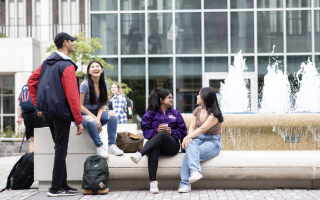 Students are seated and standing around a fountain on the UAlbany campus on a clear bright day.