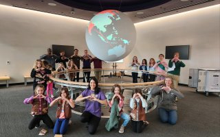 Members of the CEHC community and a group of young Girl Scouts make a heart symbol with their hands inside the ETEC "Science on a Sphere" room.