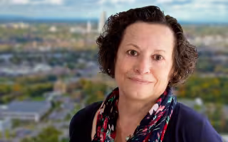 Portrait of Jean Moore wearing a navy top, red and blue flower printed scarf and gold necklace with a small green pendant. Jean has short brown hair and is smiling. The background is blurred and depicts a view of the Albany skyline.