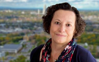 Portrait of Jean Moore wearing a navy top, red and blue flower printed scarf and gold necklace with a small green pendant. Jean has short brown hair and is smiling. The background is blurred and depicts a view of the Albany skyline.