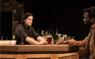 A man in a pinstripe shirt and vest stands behind a wooden bar and looks at a customer seated at the bar. A tall glass of beer sits between them.