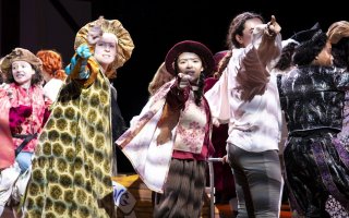 Costumed women in capes, hat and vests point toward the camera as they rehearse a play