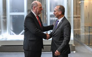 Two men in dark suits face each other and shake hands in front of a brightly illuminated enclosure holding a quantum computer.