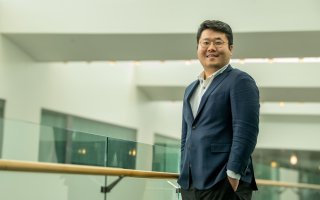 A man in glasses and a blue jacket stands, hands in pockets, near a railing in an office building