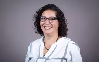 A woman with shoulder-length curly brown hair and glasses wears a striped blouse and smiles for a portrait against a gray backdrop.