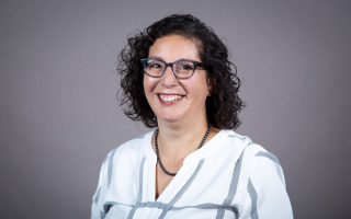 A woman with shoulder-length curly brown hair and glasses wears a striped blouse and smiles for a portrait against a gray backdrop.