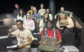 A dozen young people sit in auditorium seats with laptops, notebooks and books and pose for a photo.