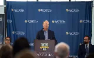 A man in a jacket stands a brown University of Rochester lectern facing an audience. 