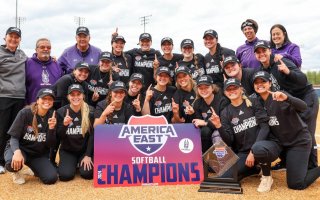 Coaches pose with members of the softball team, smiling and raising index fingers to indicate "No. 1," with the America East trophy and a sign reading "America East Softball Champions."