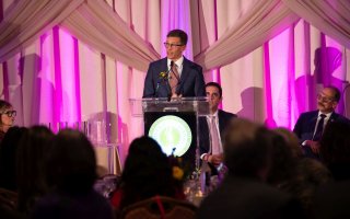 A man stands at a podium on front of an elaborate curtain, addressing a crowd at a gala