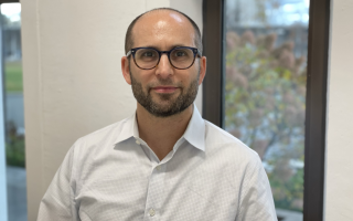 A man with close-shaved hair and glasses wears a white button-down shirt and poses for a portrait against a window.