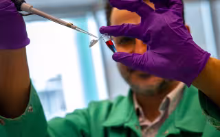 A man with brown hair and glasses holds a small plastic vial containing liquid that is purple at the bottom and red on top. He is wearing purple gloves and a green lab coat.