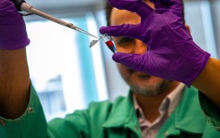 A man with brown hair and glasses holds a small plastic vial containing liquid that is purple at the bottom and red on top. He is wearing purple gloves and a green lab coat.