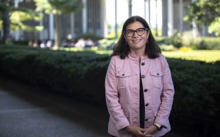 Erin Bell stands outdoors in front of a hedge, wearing a pink jacket and glasses