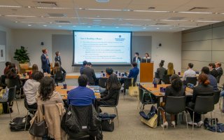 A room full of college-age students and business mentors sit at tables and view a projection screen
