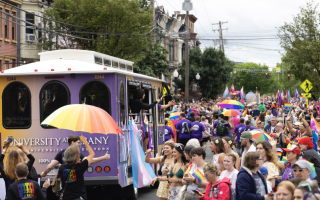 Provost Carol Kim waves to a crowd of onlookers from inside a purple and gold UAlbany trolley at the Capital Pride Parade and Festival in Albany. Throngs of people in colorful outfits and holding rainbow umbrellas fill the street.