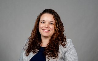Sarah Domoff poses for a portrait against a gray background wearing a light gray jacket over a navy blue shirt. Domoff is smiling and has brown curly hair. 