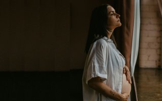 A women in an open nightshirt, hands on her pregnant belly, stares out a window.