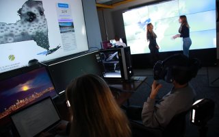 Two people sit a screens in a darkened computer lab, with one of them wearing VR goggles, as to people speak in the background.