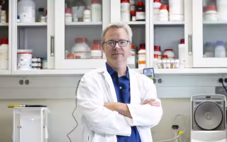 Associate Professor of Nanobioscience Ben Boivin wearing a white lab coat and blue shirt and glasses stands in his lab in front of a cabinet with clear glass panels and numerous white bottles with red caps.