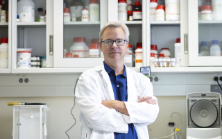 Associate Professor of Nanobioscience Ben Boivin wearing a white lab coat and blue shirt and glasses stands in his lab in front of a cabinet with clear glass panels and numerous white bottles with red caps.