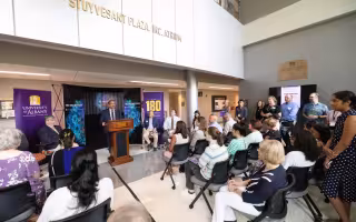 Image shows a bright sunlit room full of people listening to a man wearing a navy suit and purple tie. Many are smiling and applauding. 