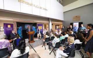 Image shows a bright sunlit room full of people listening to a man wearing a navy suit and purple tie. Many are smiling and applauding. 