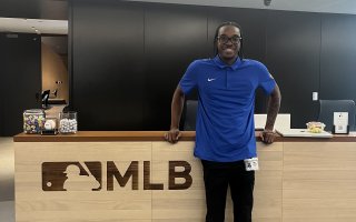 Cameron Felix stands in front of a welcome desk at MLB headquarters wearing a blue polo with his name badge attached.