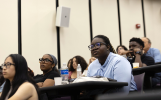 Students sit at desks in a lecture hall and look toward the front of the room.