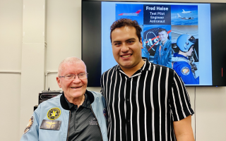A young man in a pinstripe shirt stands next to an older man in a blue space jacket as they smile for a photo. Behind them a screen reads "Fred Haise test pilot engineer astronaut"