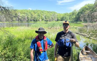 Evan Walters and another DEC intern stand in a marsh next to a canoe wearing life jackets.