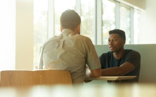 Two men talk at a table, one with his back to the camera