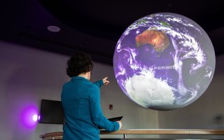 DAES Assistant Professor Aubrey Hillman points at a map of Earth's atmospheric conditions inside the Science on a Sphere room.