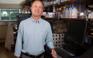 RNA Intstitute Director Andrew Berglund stands in his lab facing camera with a computer and many glass tubes in the background.