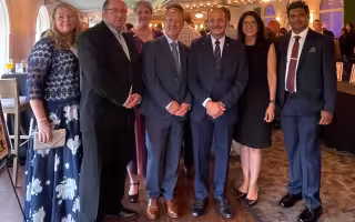 Seven people representing University at Albany leadership and the RNA Institute stand in a row, smiling for a photo at the Muscular Dystrophy Association event. Festive lighting borders the image. Left to right: Tammy Reid, John Cleary, Sheila Seery, Andy Berglund, President Havidán Rodríguez, Rosy Rodríguez, Jibin Abraham Punnoose.
