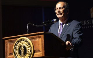 UAlbany President Havidán Rodríguez stands behind a wooden podium with the UAlbany seal and speaks into a microphone.