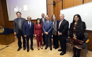 Group of seven individuals standing in front of University at Albany and AI logos, smiling in a conference room setting.