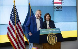 A man in a blue sport jacket and a woman in a dark business suit stand at a lectern next to an American flag. The sign on the lectern says: "Leading the Nation in Semiconductor Innovation"