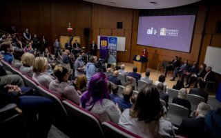 A woman in a red suit stands at a lectern in front of a packed auditorium.