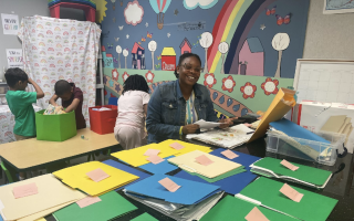 A woman in a jean jacket holds papers and smiles as she is seated at a table topped with colorful folders in a classroom with kids.