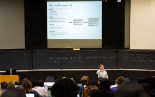 View of a classroom from the back, with students' heads visible and a professor lecturing in front of a screen with the words "Effective Management" above charts.