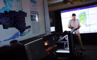 A man with an ipad looks down while a weather map of NEw York state appears in the foreground at UAlbany's XCite Lab.