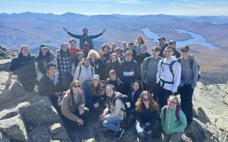 An undergraduate class stands atop Whiteface Mountain with fall foliage behind them.