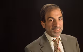 A man with short gray hair wearing a tan suit and gold tie with red stripes smiles for a portrait against a black backdrop.