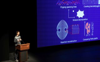 A man speaks from a podium next to a projection of mathmatical images.