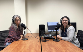 Two women wearing headphones sit in front of microphones at a table next to a computer and recording equipment.